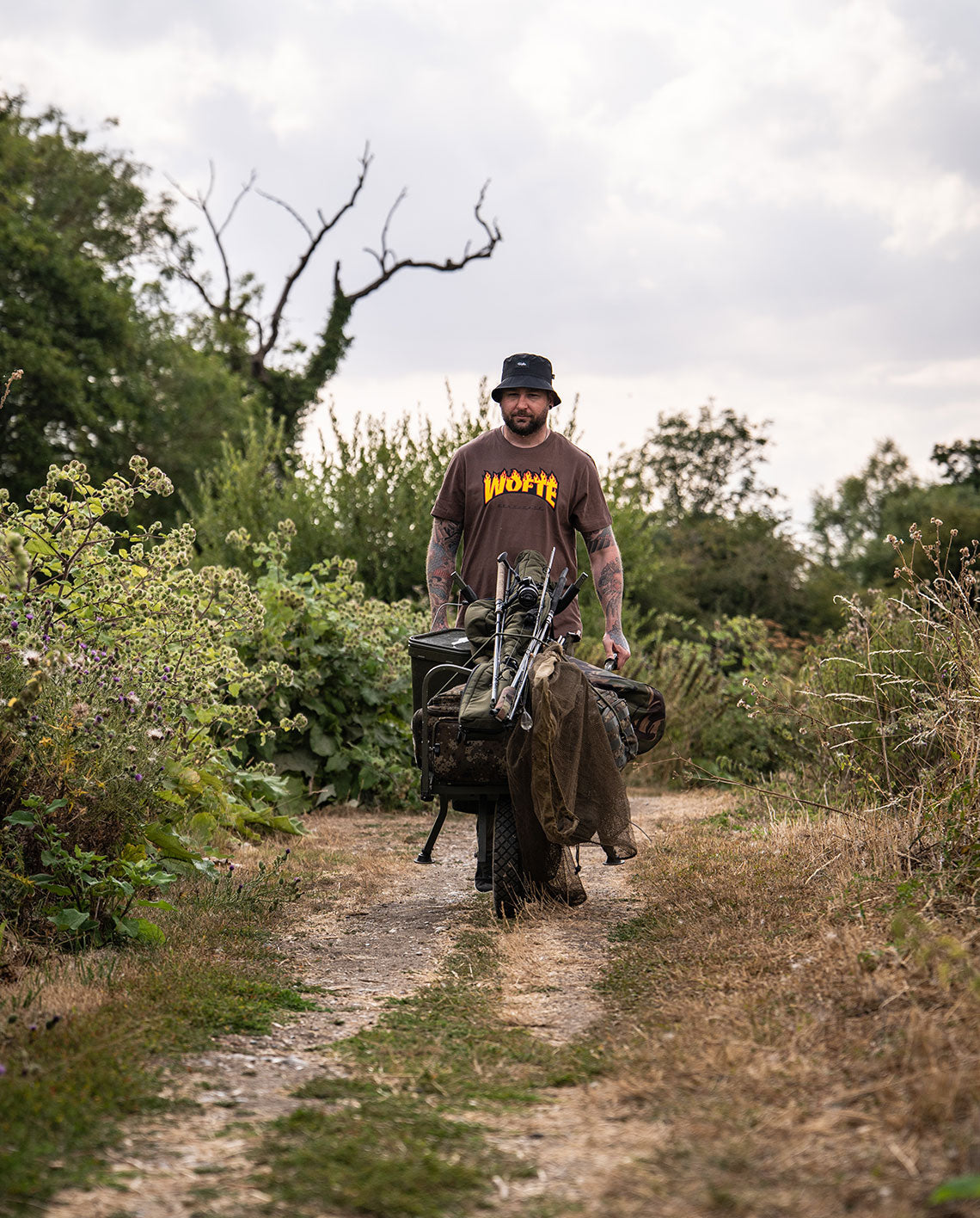 An image of a man pushing his carp porter barrow wearing a Wofte Syndicate t-shirt in light brown. 